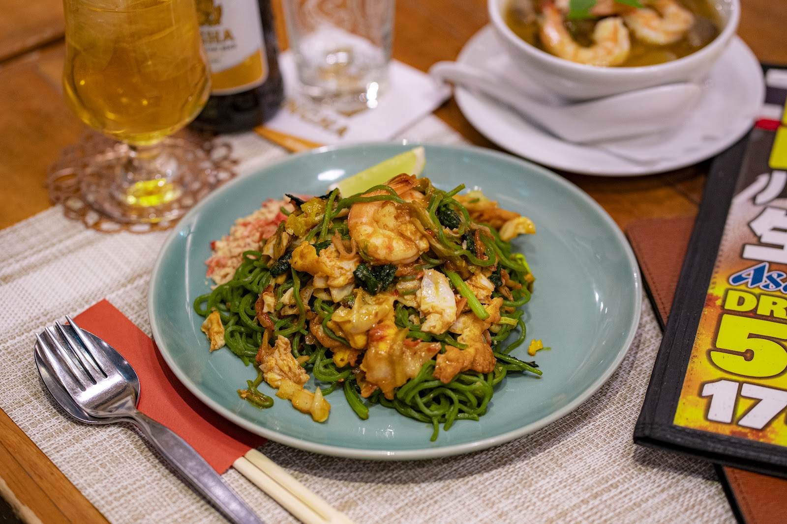 青緑の皿に盛った緑色の麺の炒め物とえび、ライムの一切れ。左にビールのグラス、奥にえびのスープの器。