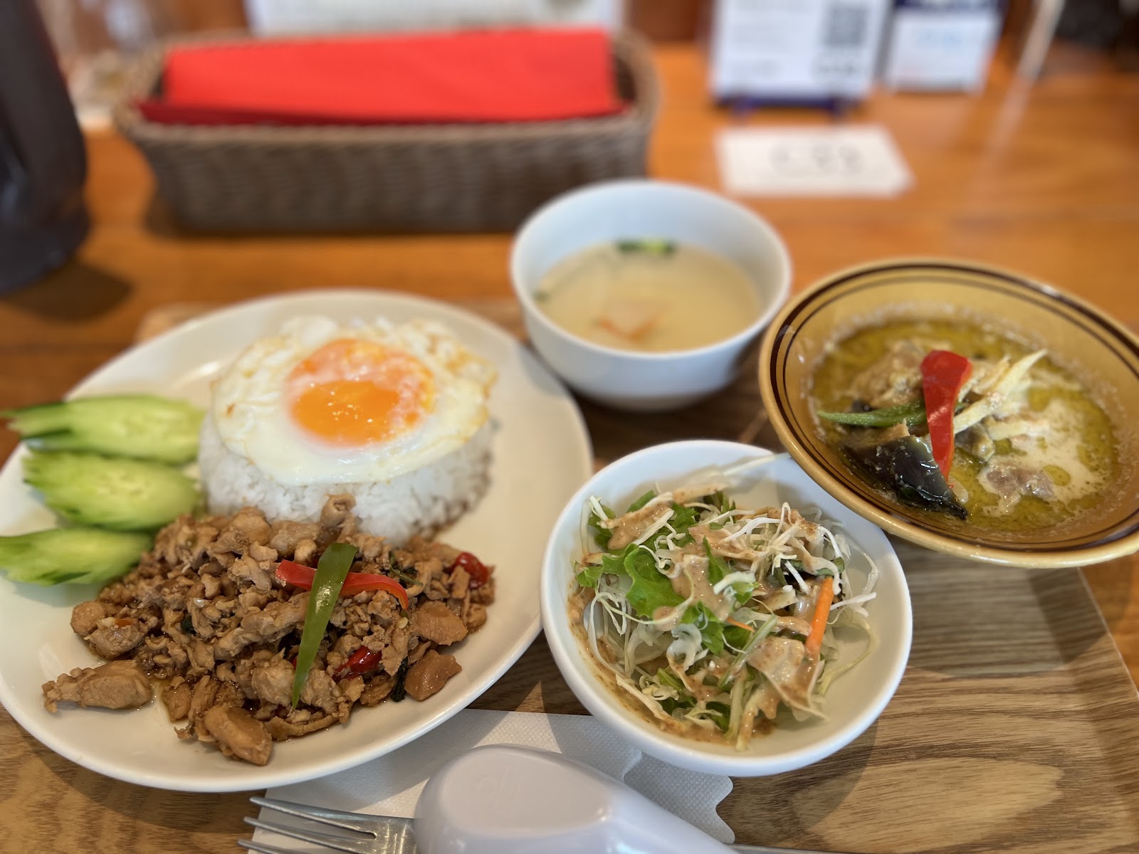 木のトレーにのった定食。目玉焼きをのせたガパオライス、きゅうり、透明なスープ、千切りキャベツのサラダ、緑色のカレーの器。