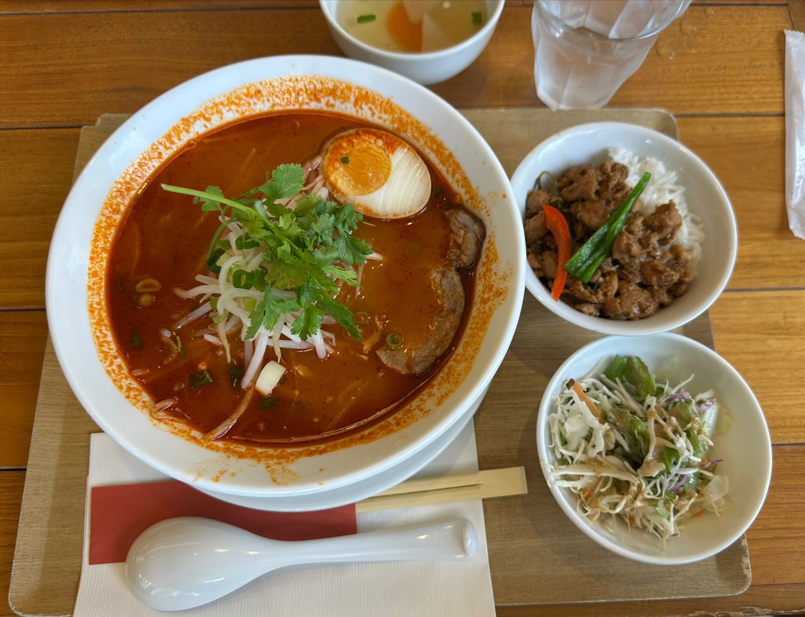 赤いスープの麺の丼。パクチー、もやし、半分の味付け卵。横に鶏ひき肉をのせたご飯の小鉢とサラダ。