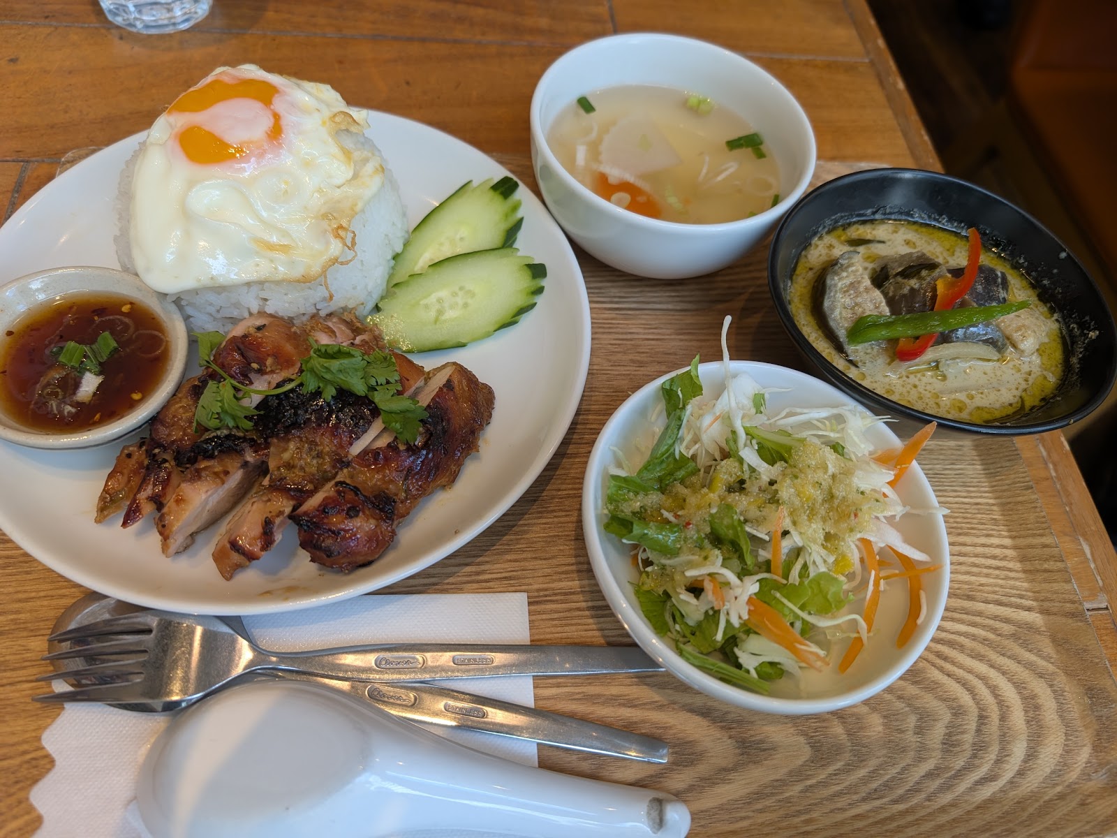 焼いた鶏もも肉とパクチー、目玉焼きをのせたご飯、たれの小皿。横にスープ、サラダ、黒い器のカレー。